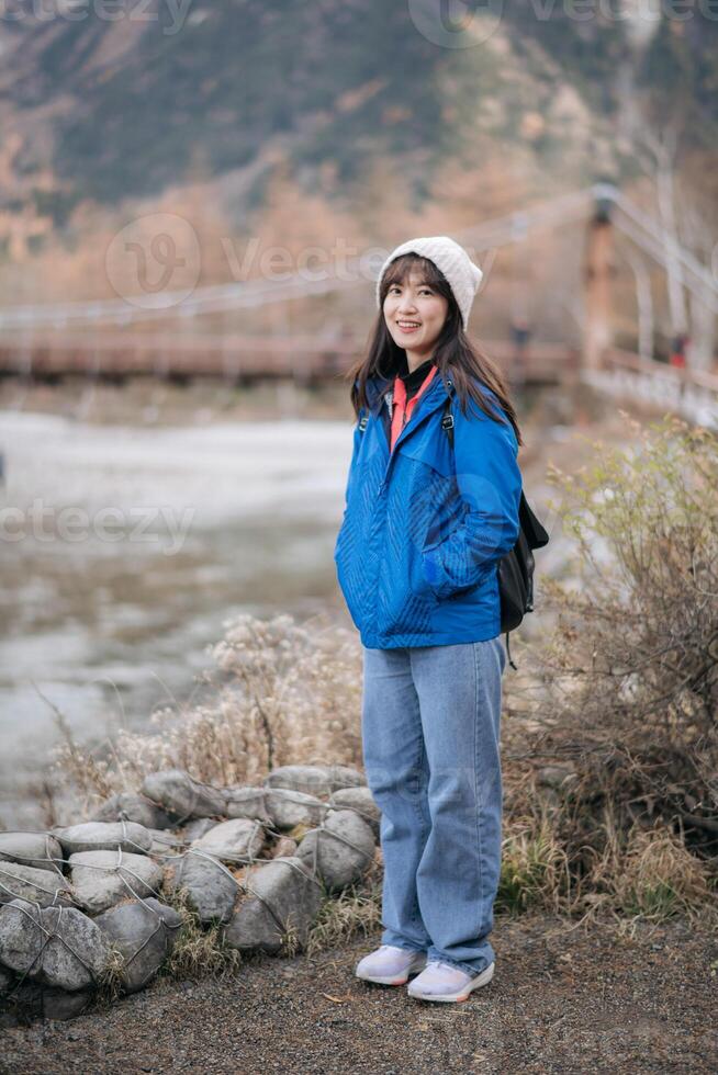 Southeast asian woman enjoys travel concept standing near river with bridge in background wearing blue jacket and white beanie expressing happiness in nature photo
