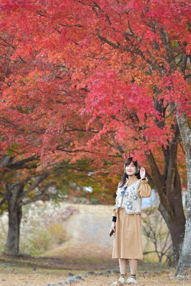 Southeast asian woman stands peacefully under vibrant red autumn leaves wearing beige dress and patterned vest, enjoying serene natural beauty of season photo