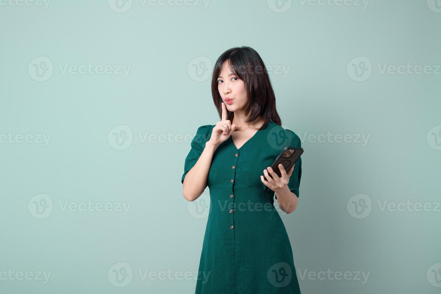 Southeast asian woman holds mobile phone and gestures to be quiet while using internet application in green dress against plain background with calm expression photo