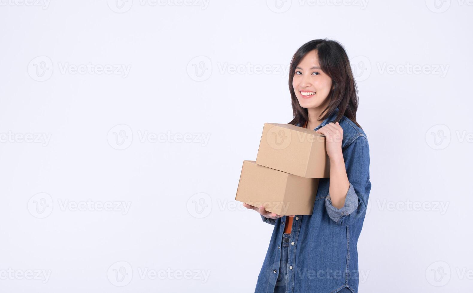 Southeast Asian woman smiling while shopping online holds two cardboard boxes in casual denim outfit against white background showing happiness and convenience photo