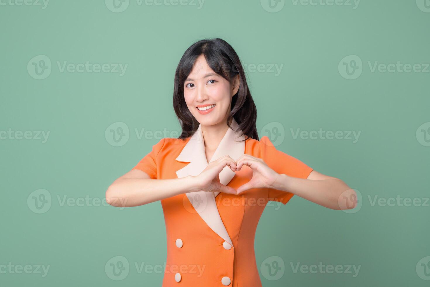 Southeast asian woman in orange dress smiling and making heart shape with hands against green background, expressing happiness and positivity in studio portrait photo