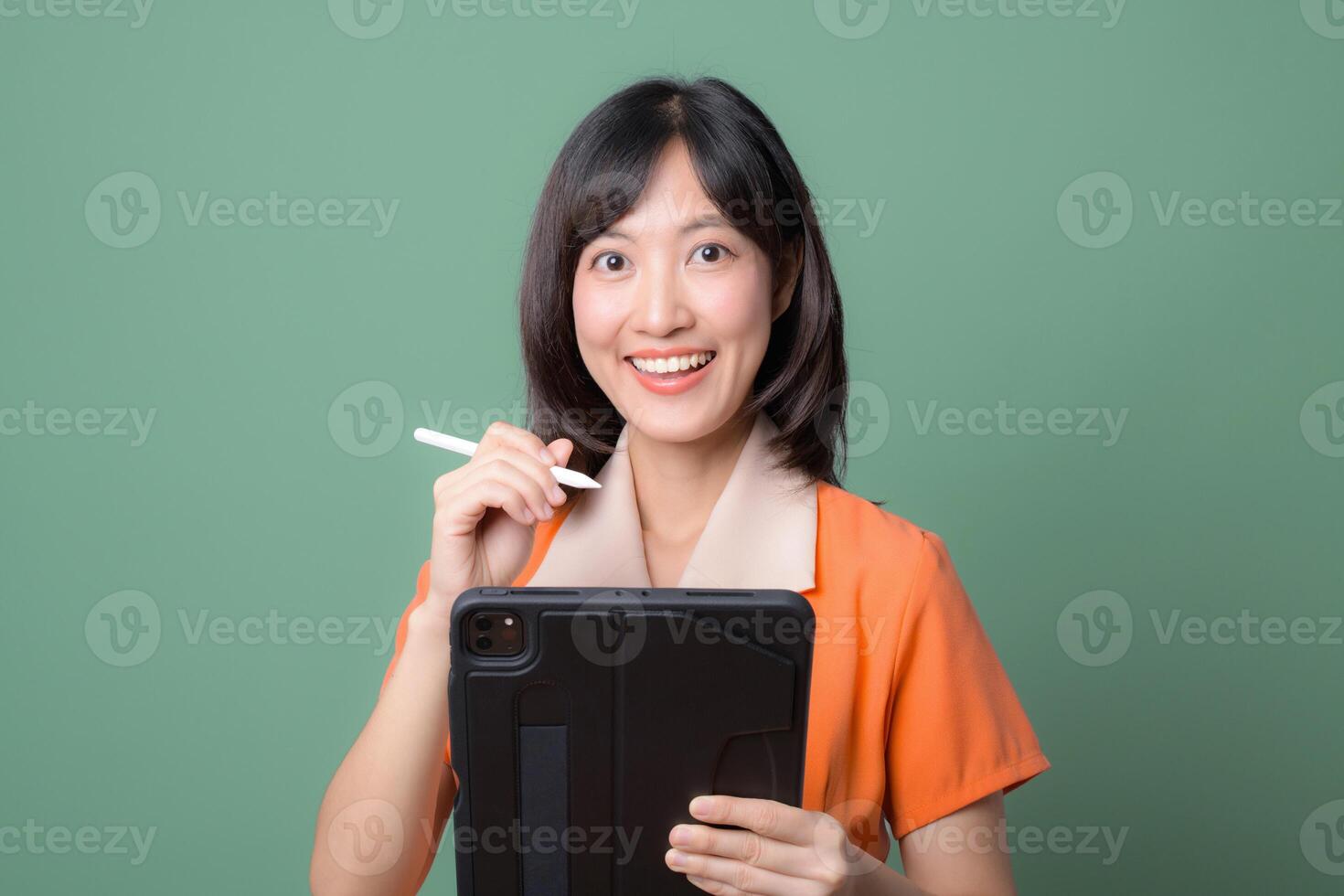Southeast asian woman in orange shirt smiling holding digital tablet and stylus against green background showing cheerful expression photo