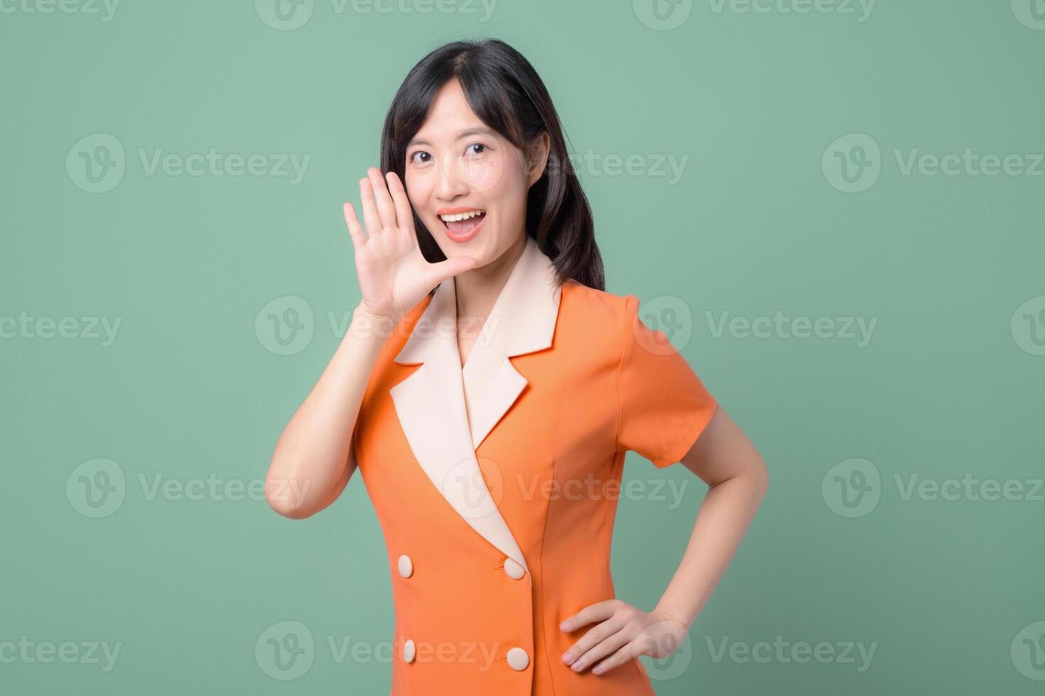 Southeast asian woman in orange dress smiling and holding hand near mouth as if calling out or sharing secret against green background cheerful expression photo