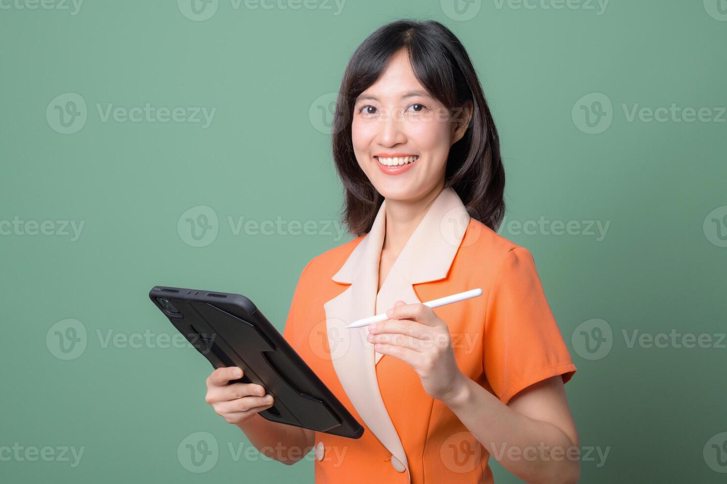 Southeast asian woman smiling while using internet on digital tablet with stylus pen, standing against green background, professional and confident expression photo