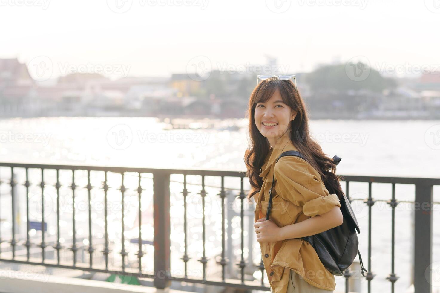 Southeast asian woman travel concept shows smiling woman with long hair wearing brown jacket and black backpack standing near waterfront railing in soft sunlight photo