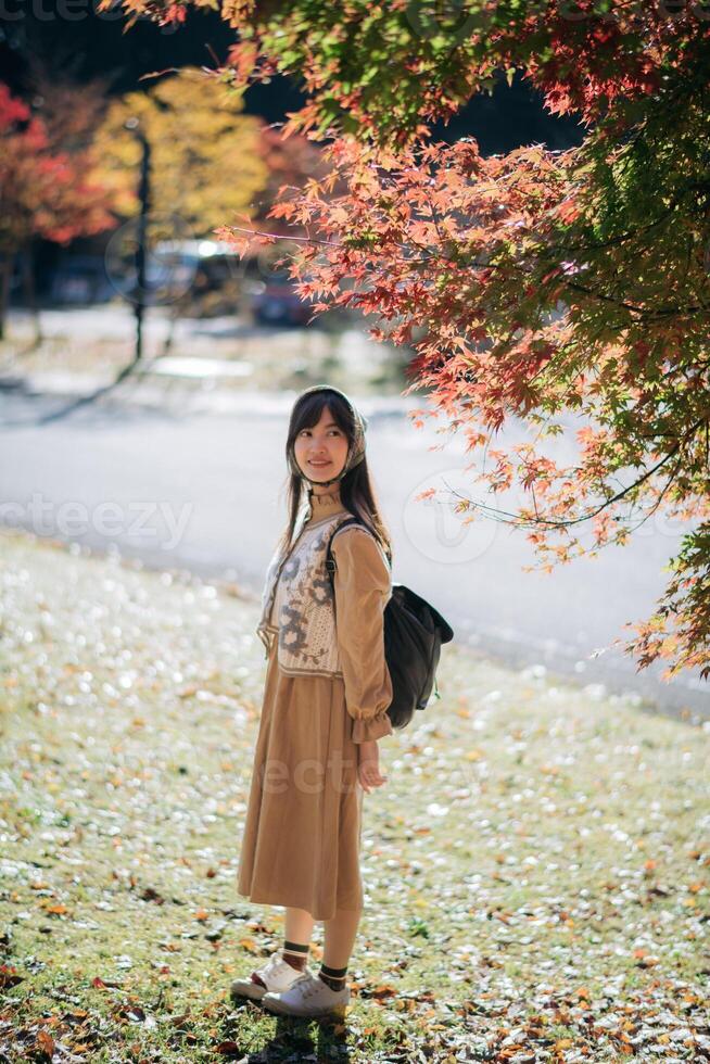 Southeast asian woman enjoys travel concept in autumn park wearing brown dress and backpack under colorful leaves with peaceful and happy expression on sunny day photo
