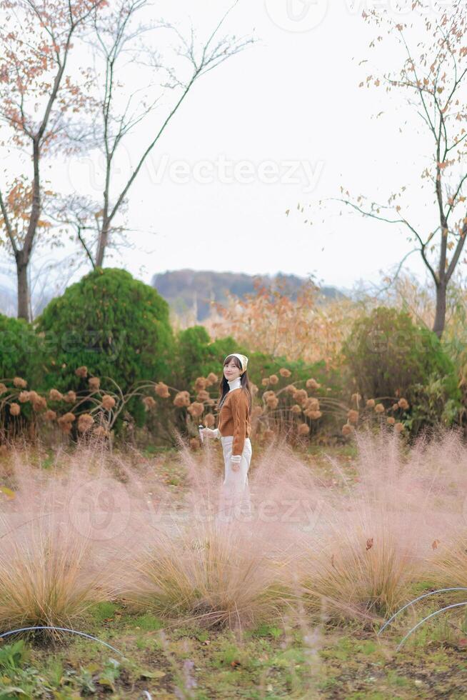 Southeast asian woman stands peacefully in field of soft pink grass with autumn trees and green bushes around her creating calm and serene outdoor scene photo