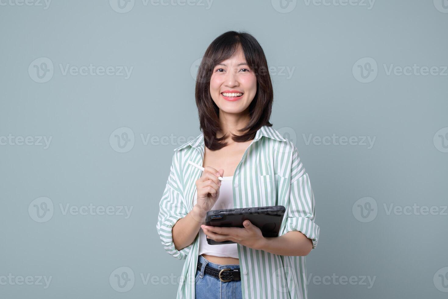Southeast Asian woman smiling while using internet on digital tablet, casual outfit, standing against plain background, cheerful and confident expression, modern lifestyle photo