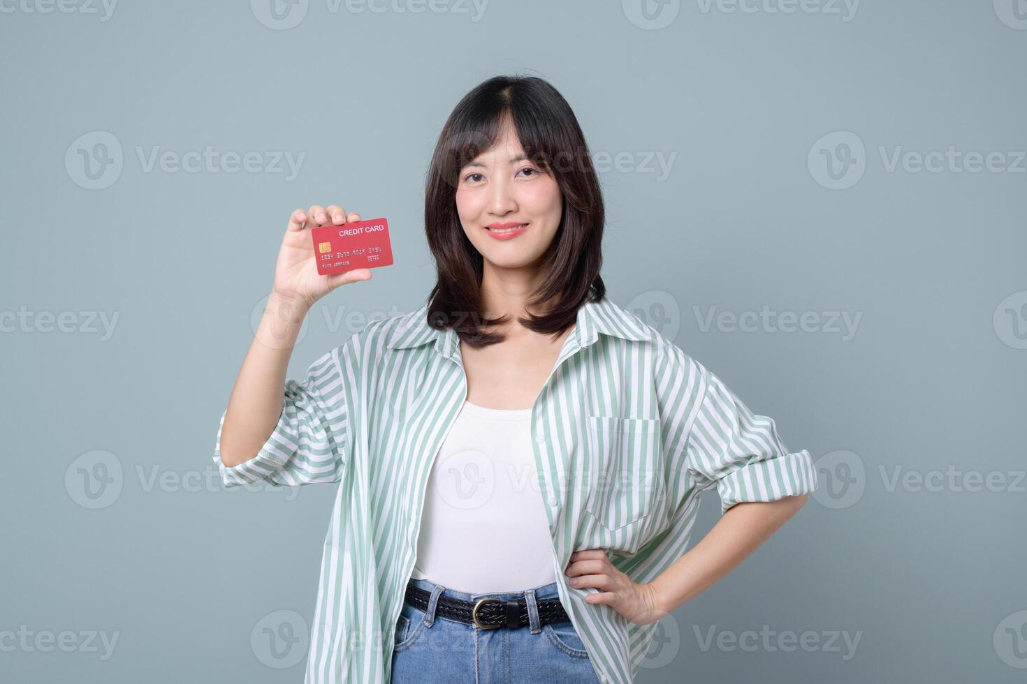 Smiling asian woman holding red credit card with happy expression, casual outfit, standing against green background photo