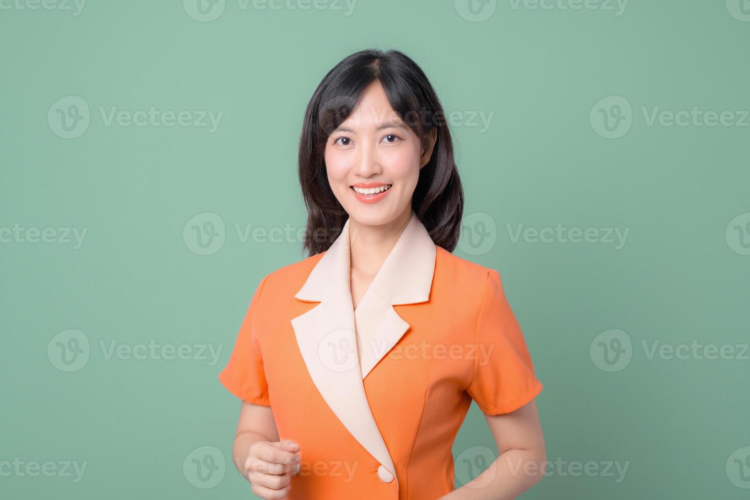 Southeast asian woman with long black hair wearing orange blazer with cream collar stands smiling confidently against plain green background in studio portrait photo