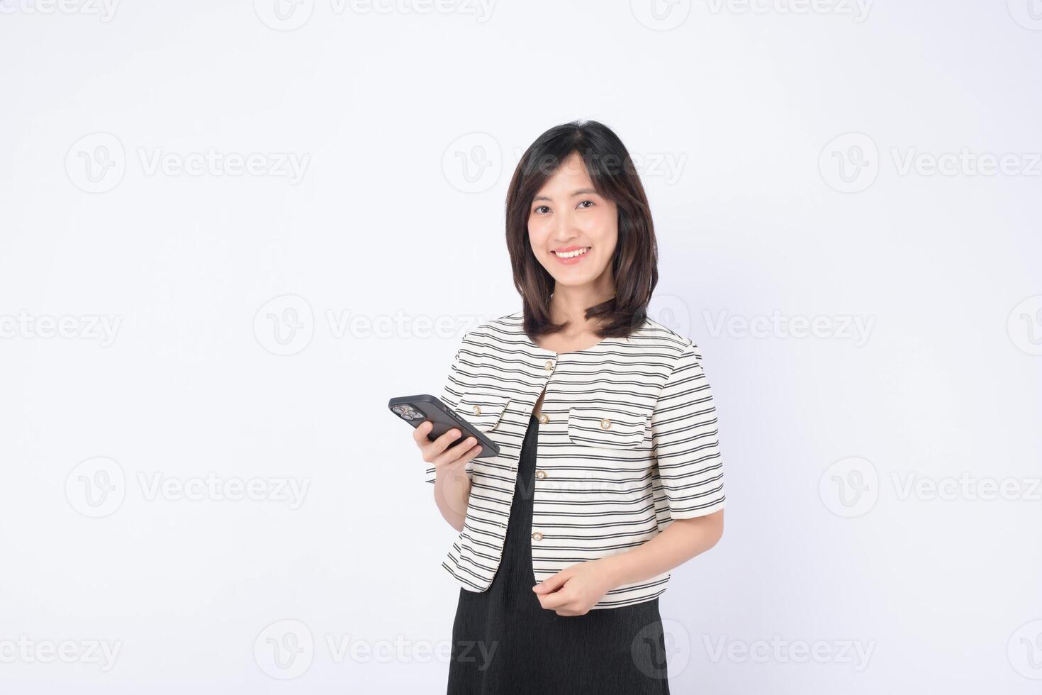 Southeast asian woman smiling holding smartphone in hand wearing striped jacket and black dress standing against white background showing confidence and happiness photo
