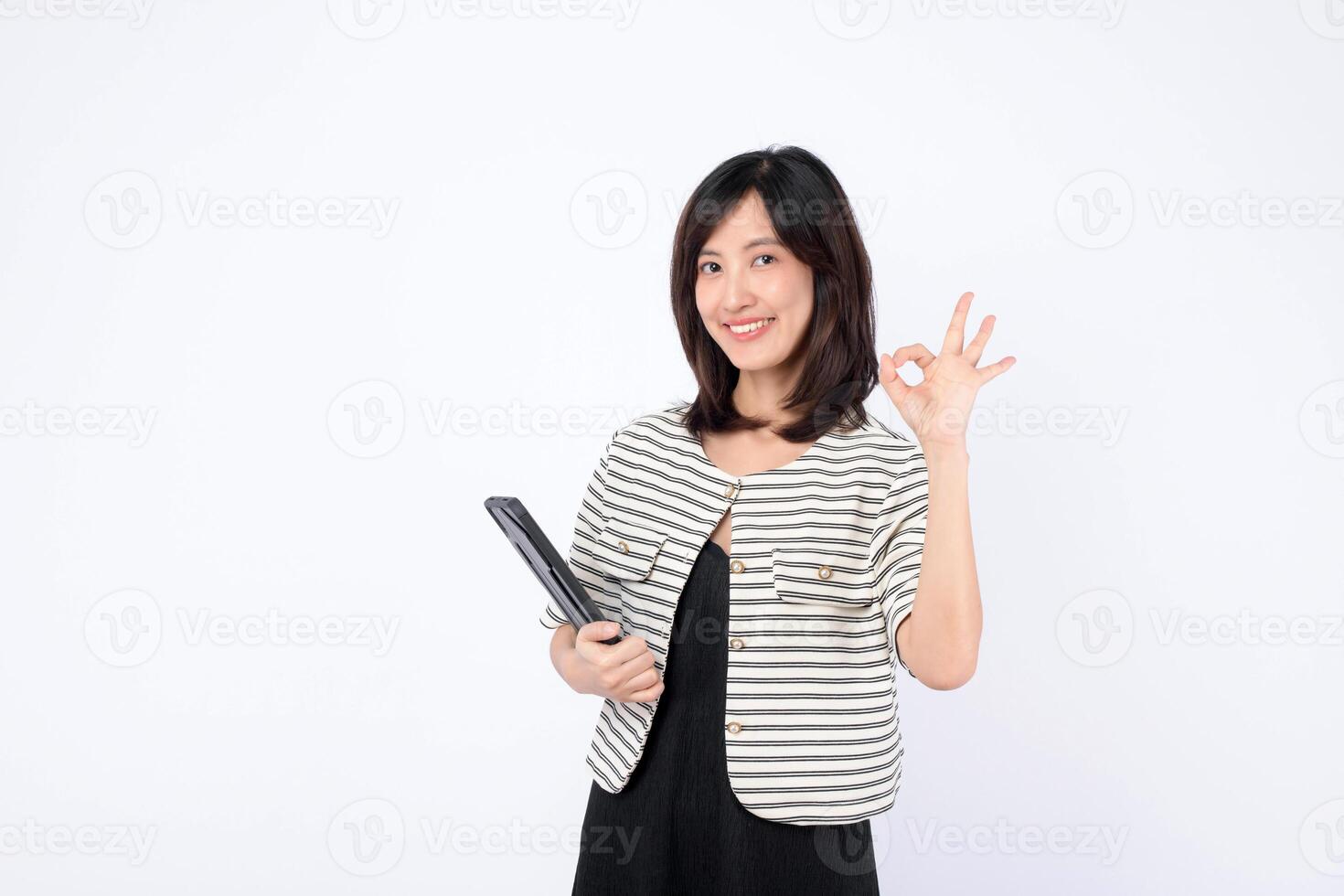 Southeast Asian woman smiling and holding tablet while making OK hand gesture, standing against white background, expressing confidence and positivity photo