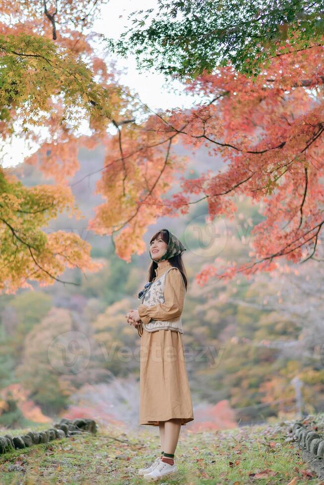 Southeast Asian woman enjoys travel in autumn forest with colorful leaves, wearing brown dress and smiling peacefully in nature, surrounded by vibrant foliage photo