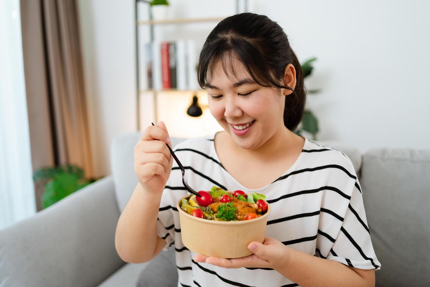 Happy young Asian woman eating vegetable salad in paper cup for health and weight loss photo