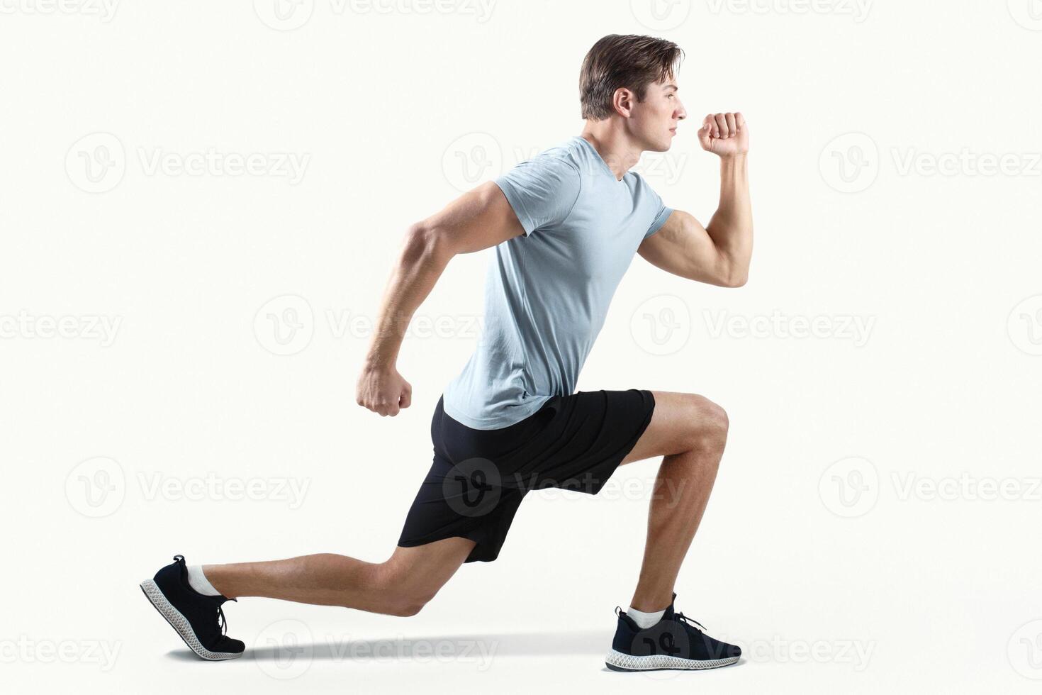 Side view of strong millennial guy in sportswear doing lunges on light studio background. Determined athletic man exercising, stretching his legs or warming up before strength training, full length photo