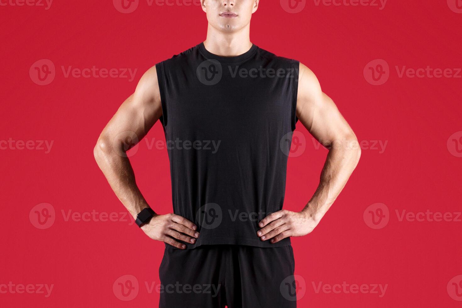 Full length portrait of determined male fitness model looking at camera on red studio background. Serious millennial athlete in black sportswear posing with hands on hips. Professional sports concept photo