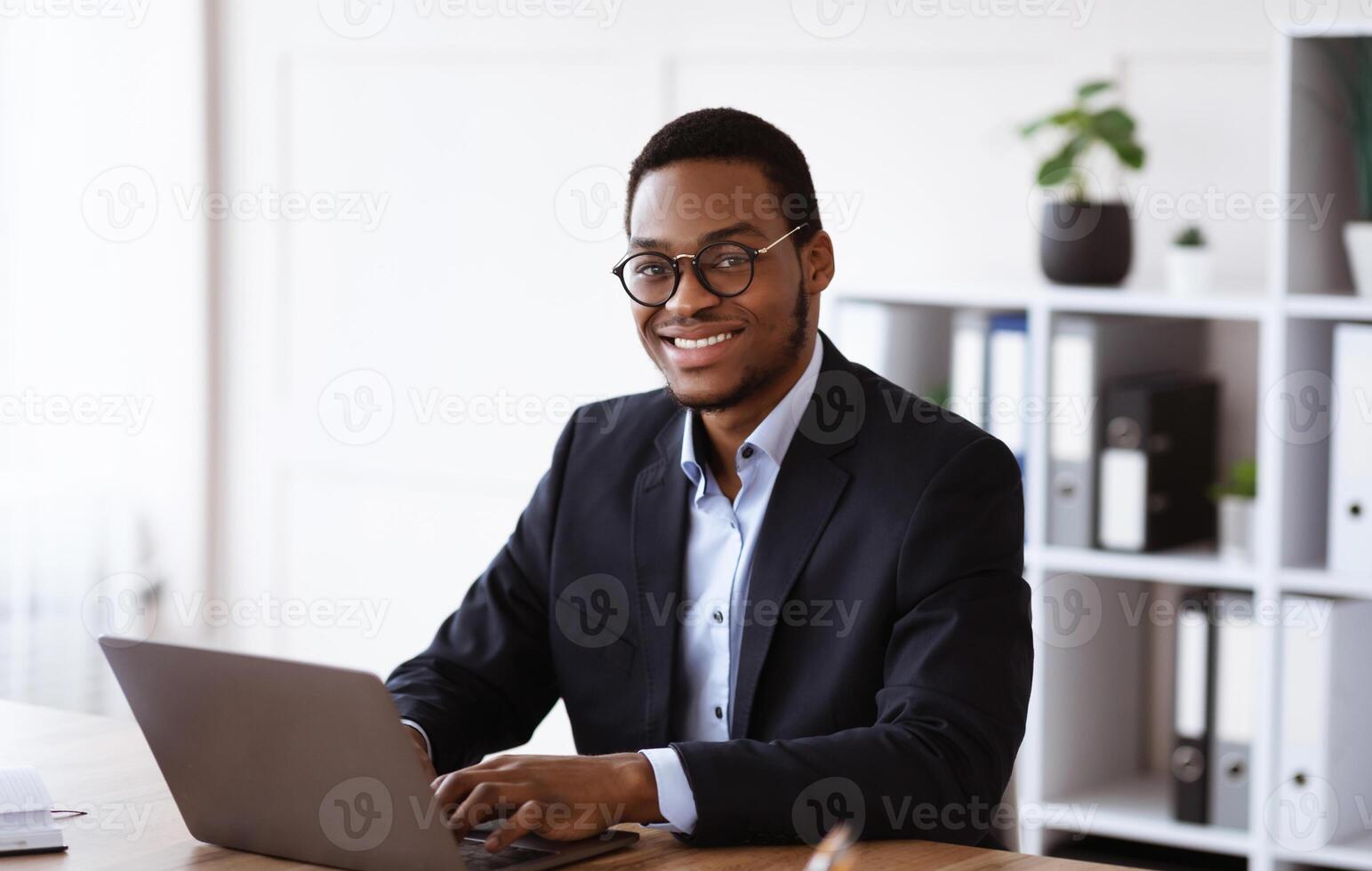 Cheerful african american businessman working with modern laptop in office, copy space. Happy young black manager in suit and glasses working online, typing emails or chatting with clients photo