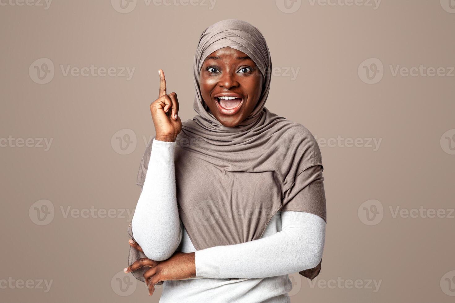 Excited Black Islamic Lady In Headscarf Having Idea, Pointing Finger Up, Emotional Africam Muslim Woman Having Inspirational Moment While Standing Over Light Background In Studio, Copy Space photo