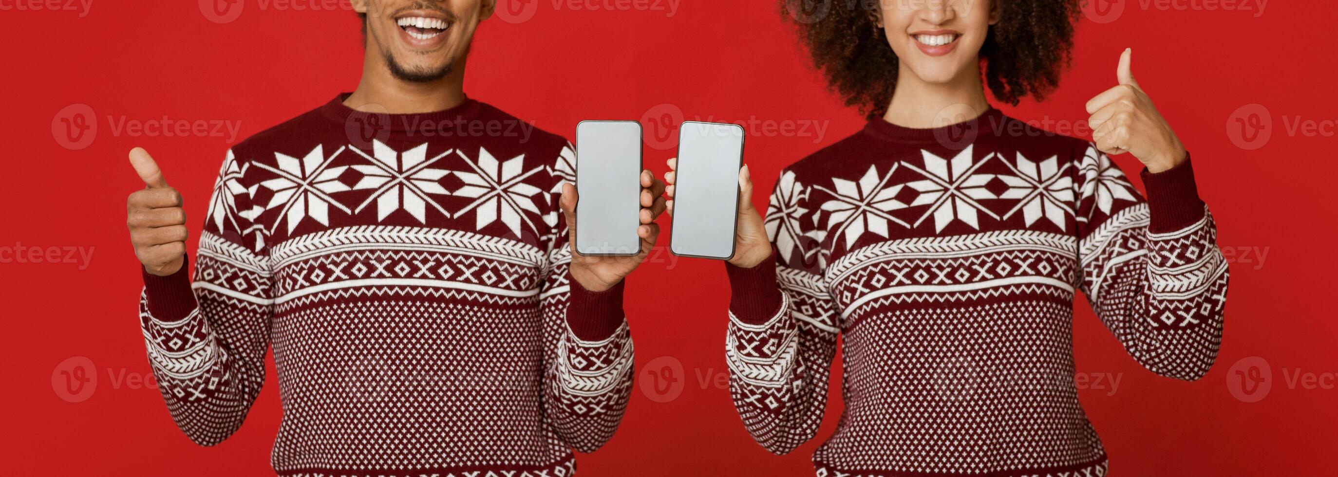 Family look in New Year and Christmas patterned sweaters. Smiling african american man and woman show thumbs up and smartphones with blank screens, isolated on red background, panorama, cropped photo