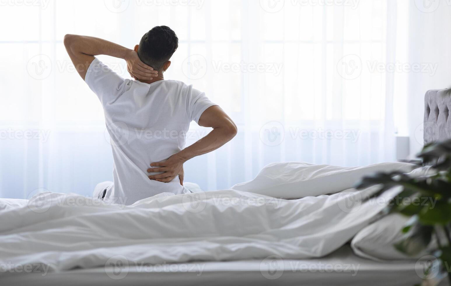Unrecognizable Young Man Waking Up In Morning With Neck And Back Pain, Millennial Male Having Backache, Massaging Inflamed Aching Zones While Sitting On Bed At Home, Rear View With Copy Space photo