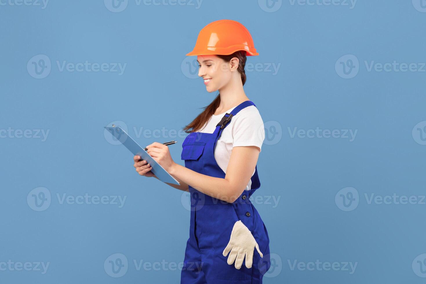 Smiling young handywoman in coveralls and hardhat writing notes to clipboard, making list of construction materials and tools for house renovation, standing over blue studio background, copy space photo