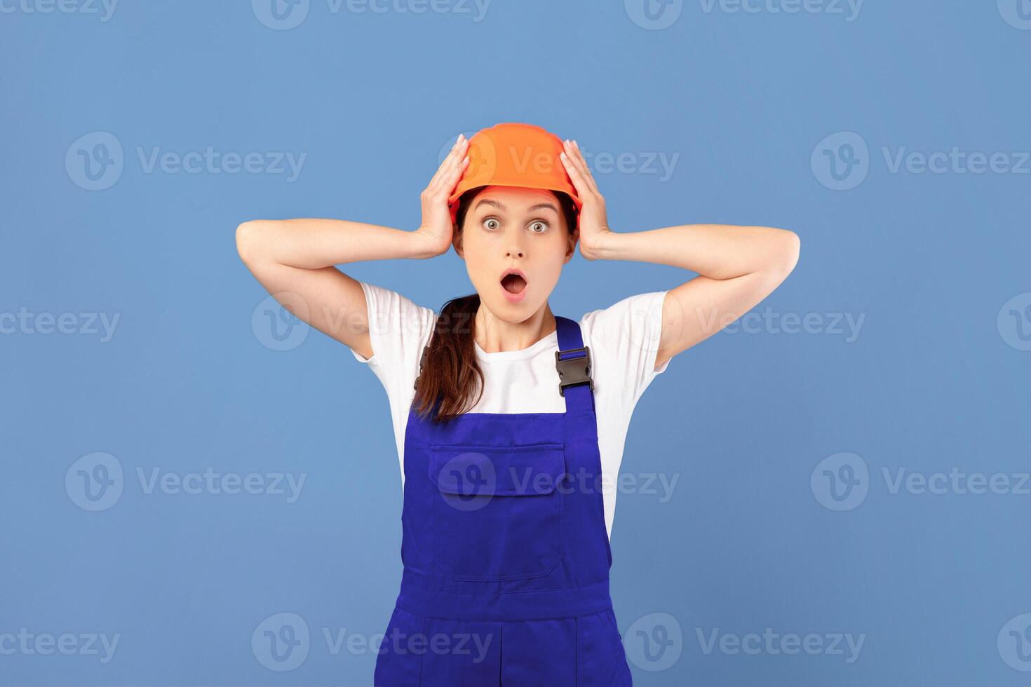 Shocked handywoman in protective helmet and coveralls touching head in excitement, amazed woman standing with opened mouth over blue background, emotionally reacting to repair issues, copy space photo