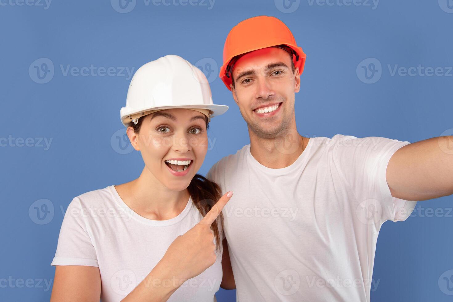 Repair Home Concept. Close up of young couple in protective helmets taking selfie together during renovation, cheerful man and woman in hard hats smiling at camera over blue background, free space photo