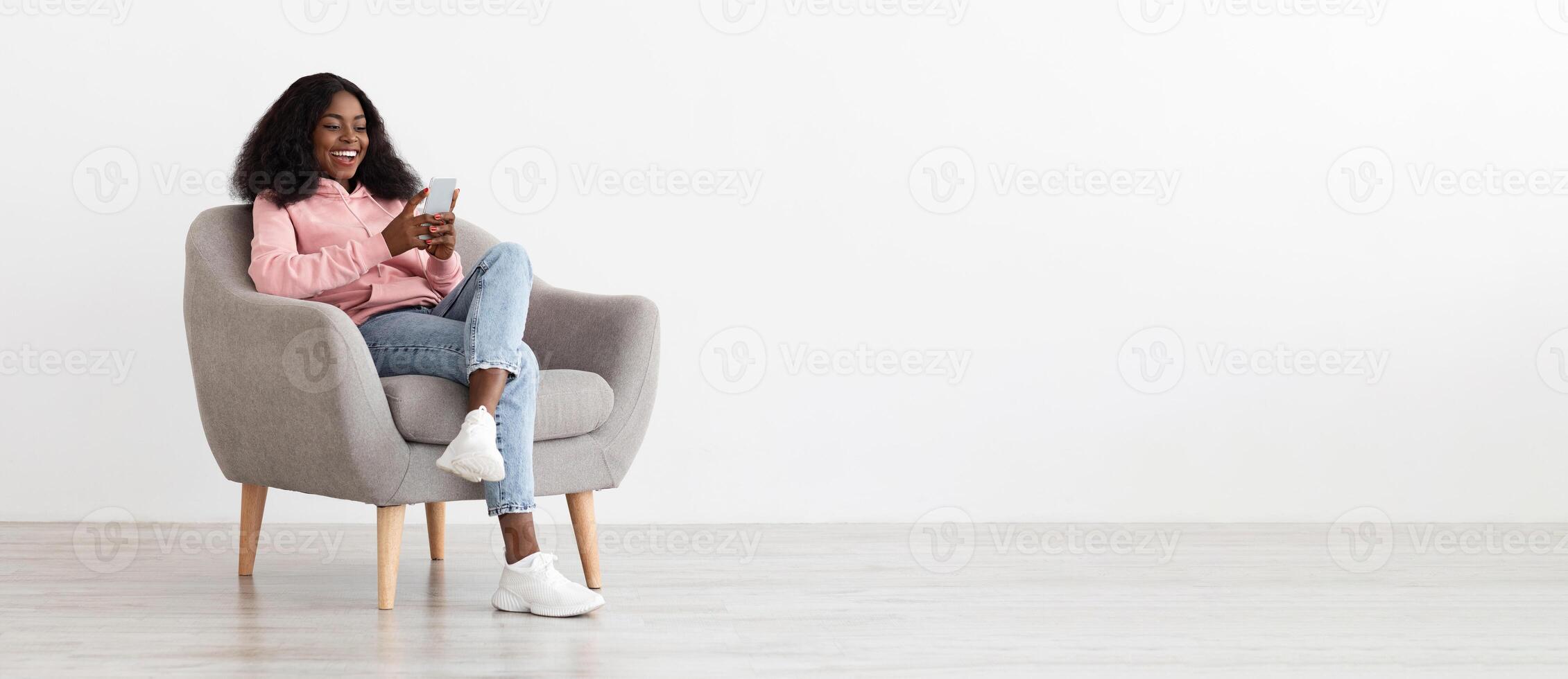 Relaxed pretty young black woman sitting in arm chair with cellphone at home, checking social media, empty white wall background, panorama with copy space. Gadgets addiction for millennials concept photo