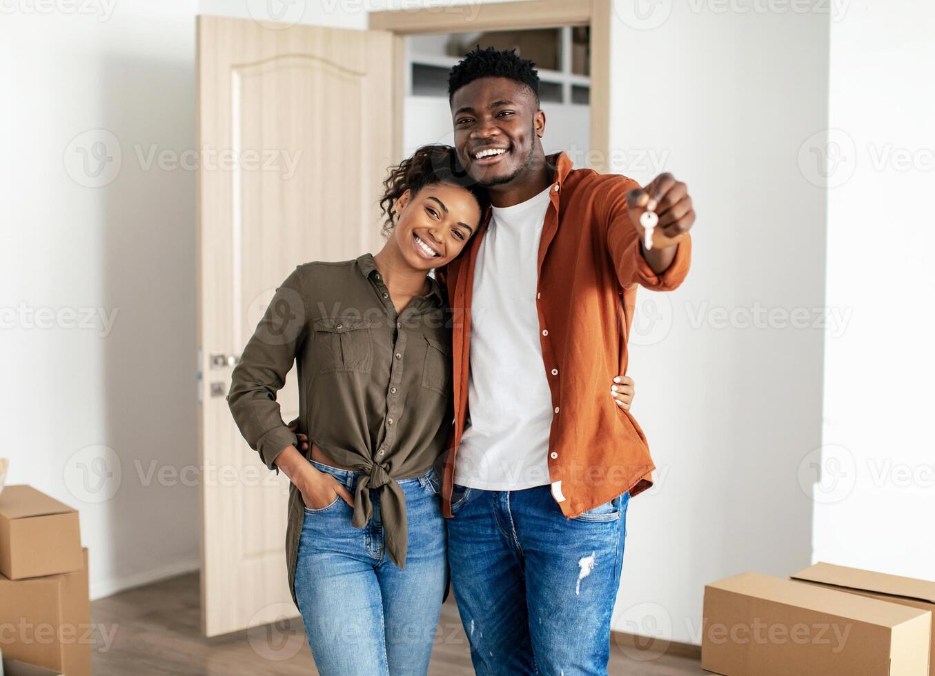 Family Housing. African American Millennial Couple Showing Key Posing Embracing In New House Among Moving Boxes. Real Estate Buyers Standing At Their Own Home, Smiling To Camera photo