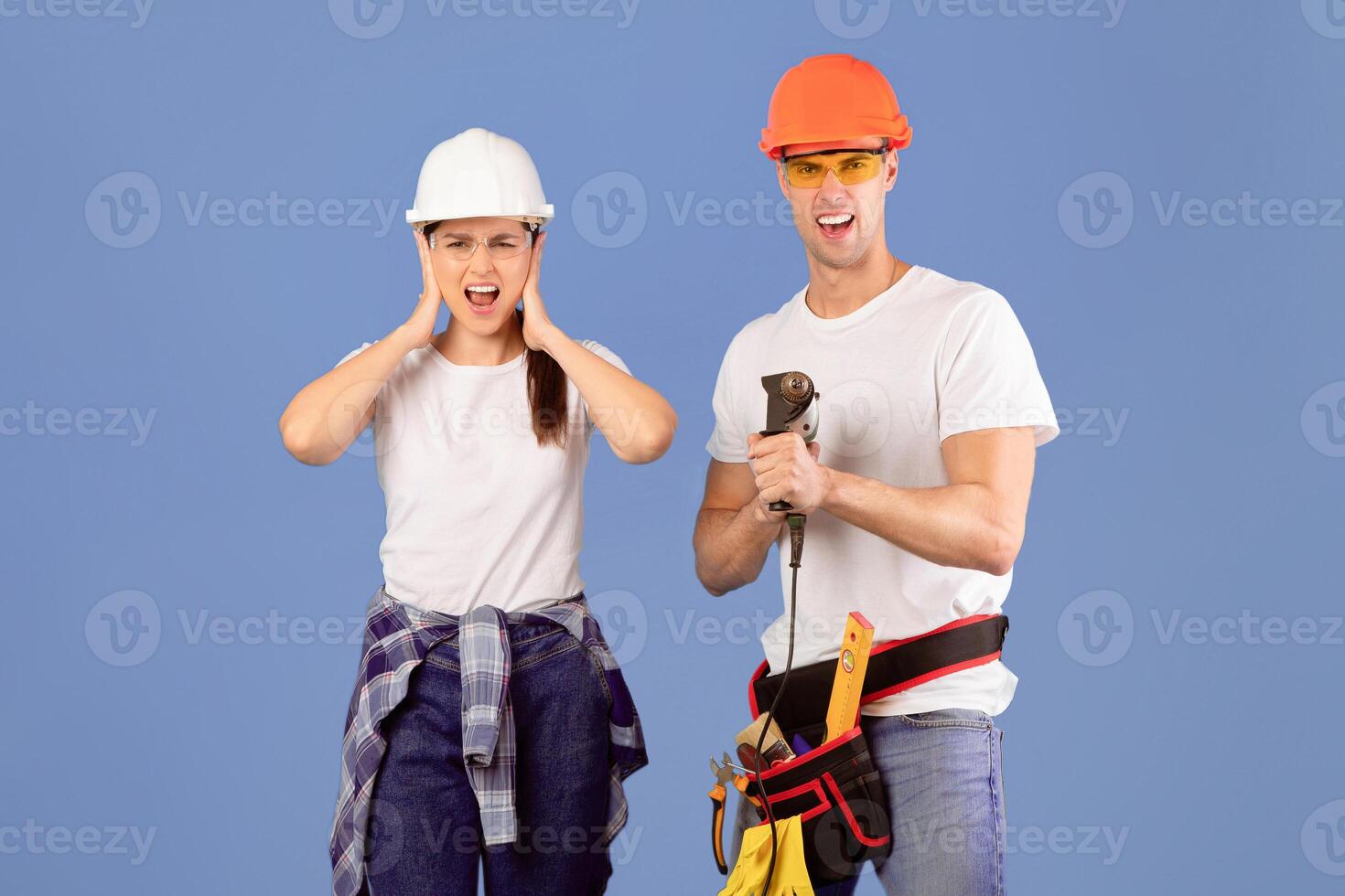 Crazy Repairment. Funny Couple In Hard Hats And Protective Glasses Posing With Construction Tools Over Blue Background, Man Aiming Drill At Camera And Woman Covering Ears, Can Not Stand Sound photo