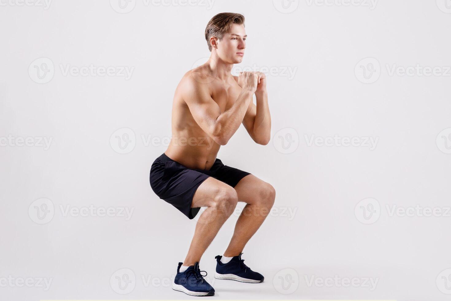 Motivated young sportsman with bare chest warming up before training, doing squats on light studio background, full length. Focused millennial athlete with naked torso doing cardio aerobic exercises photo