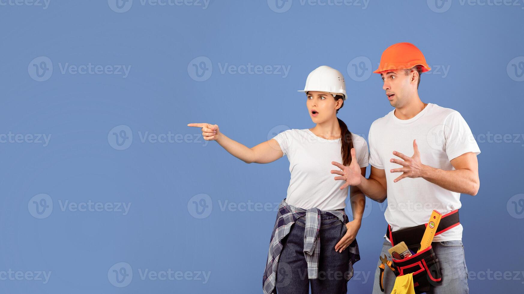 Shocked Young Couple In Hard Hats Pointing Aside At Copy Space Over Blue Background, Emotional Man And Woman In Protective Helmets And Casual Clothes Making House Renovation Together, Panorama photo