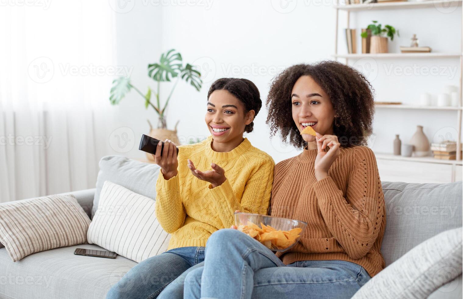 Great movie and relax at home in spare time. Funny millennial african american and mixed race females watching TV with remote control in hand and eating chips, sits on couch in living room interior photo