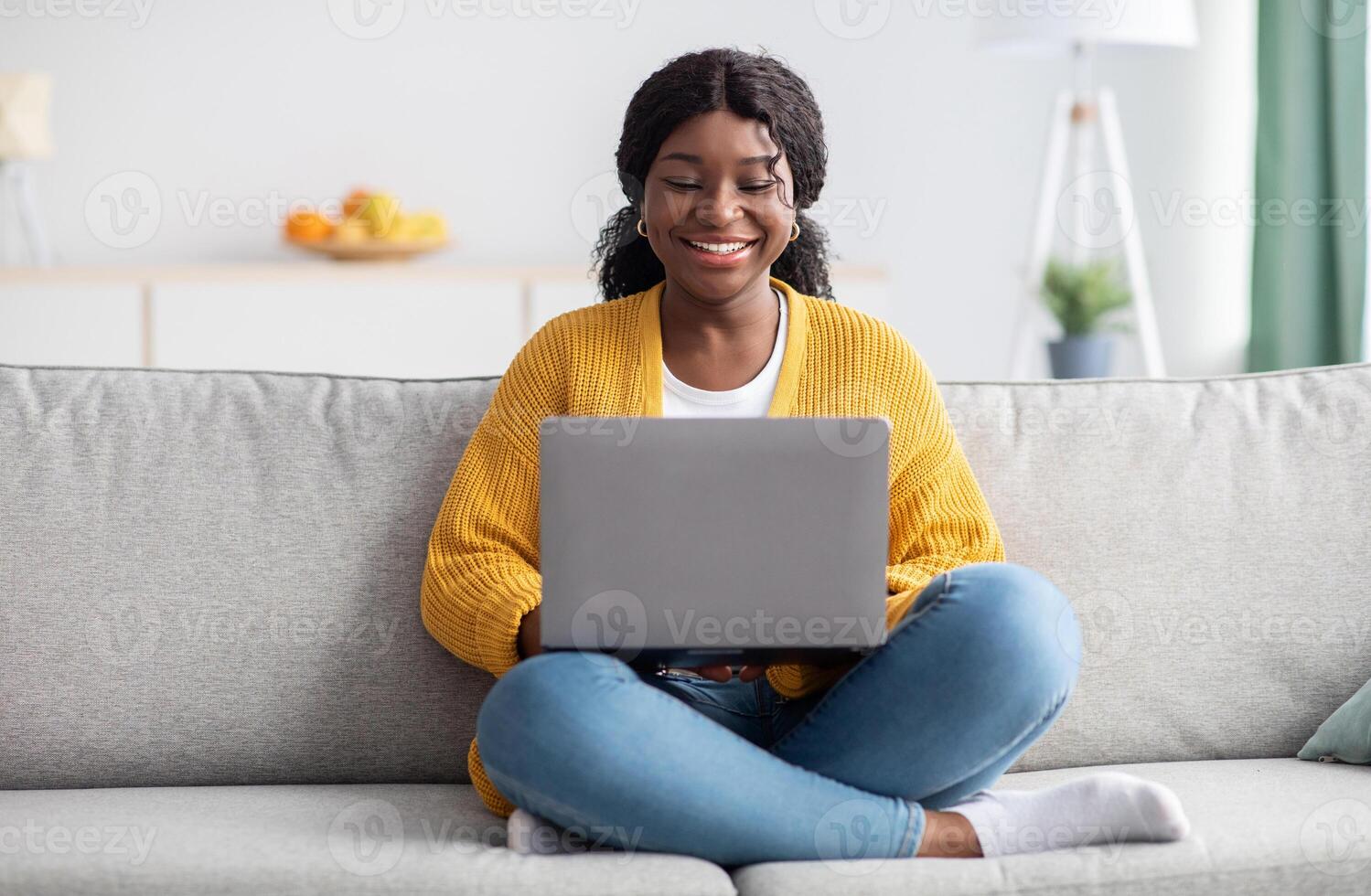 Smiling young black woman in knitted sweater freelancer working from home, sitting on couch, using laptop, copy space. Relaxed african american lady enjoying her weekend, surfing on internet on laptop photo