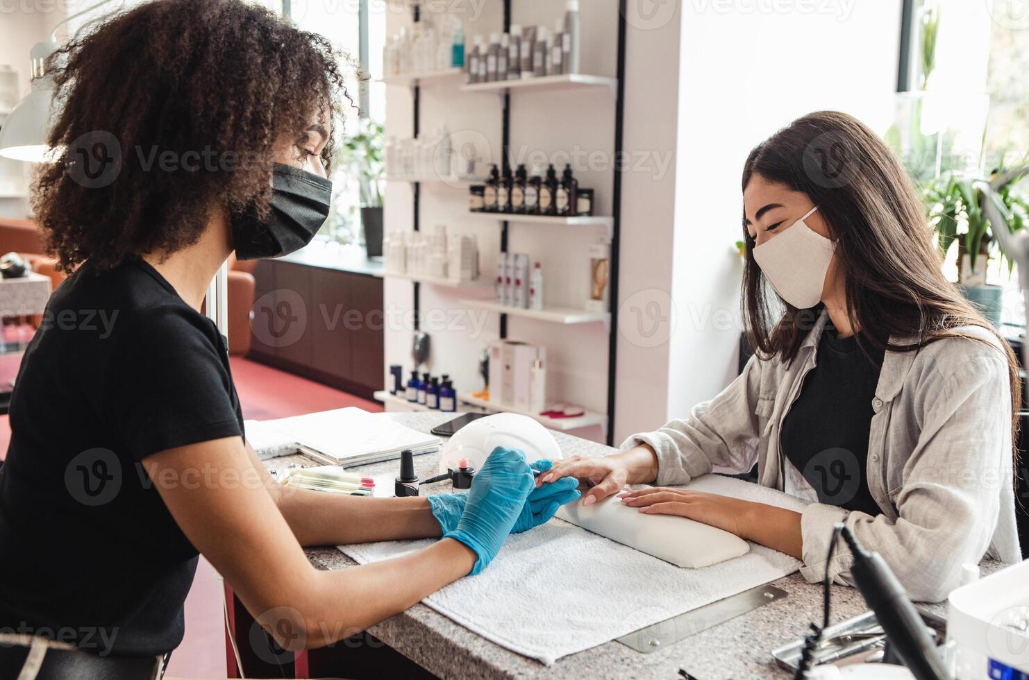 Returning to work after quarantine and gossip at nail salon. African american woman in protective mask files nails with electric device to smiling asian lady client in studio interior photo