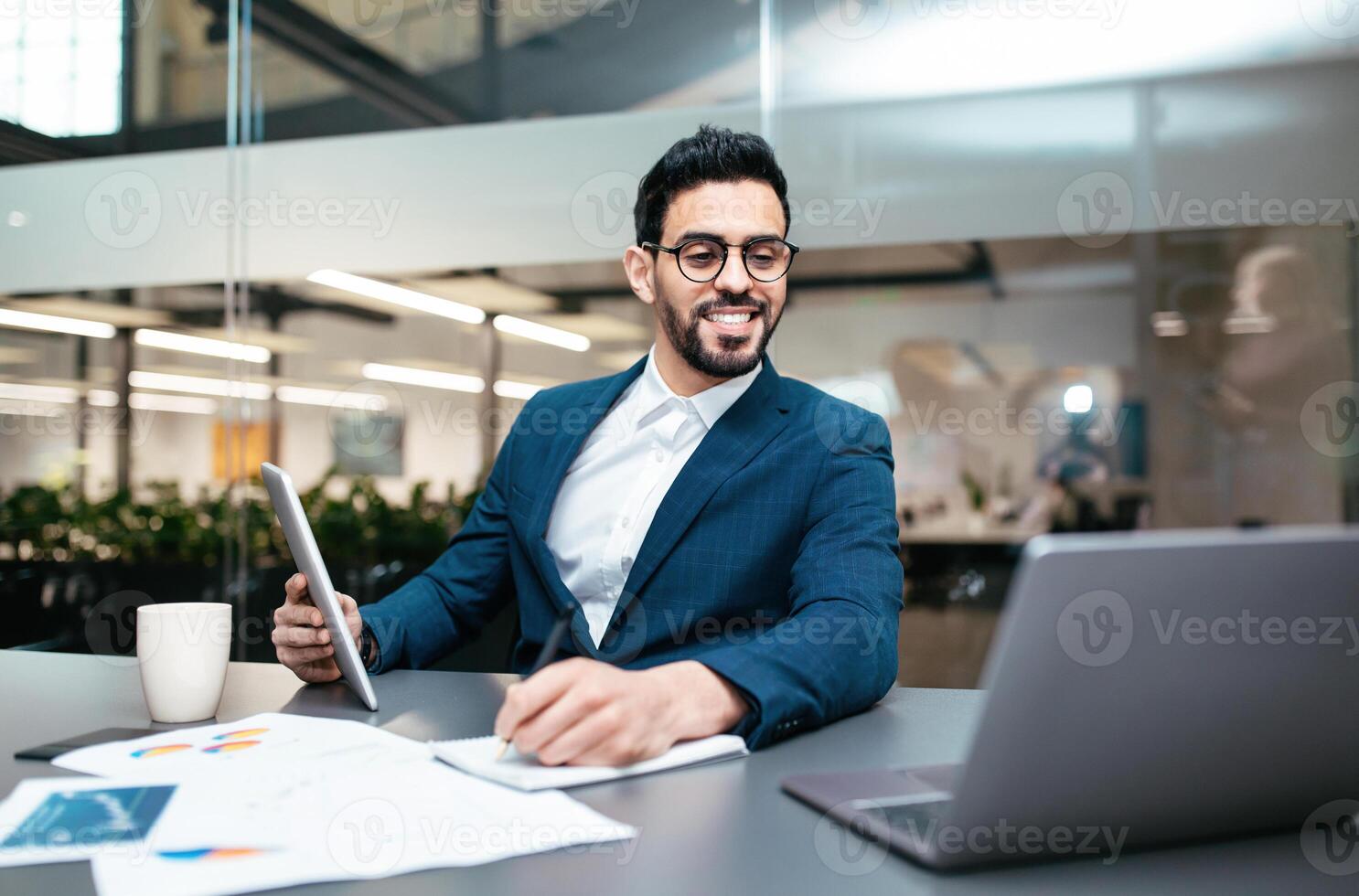 Happy attractive young islamic male with beard in glasses, suit sits at table with computer and tablet in office interior. Professional working remotely, business and modern technology, new normal photo