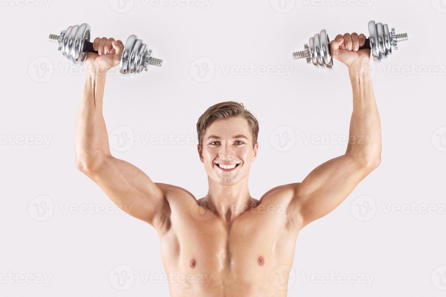 Portrait of positive young athlete lifting dumbbells above his head and smiling on light studio background. Strong bodybuilder with six pack abs working out his biceps, pumping iron photo