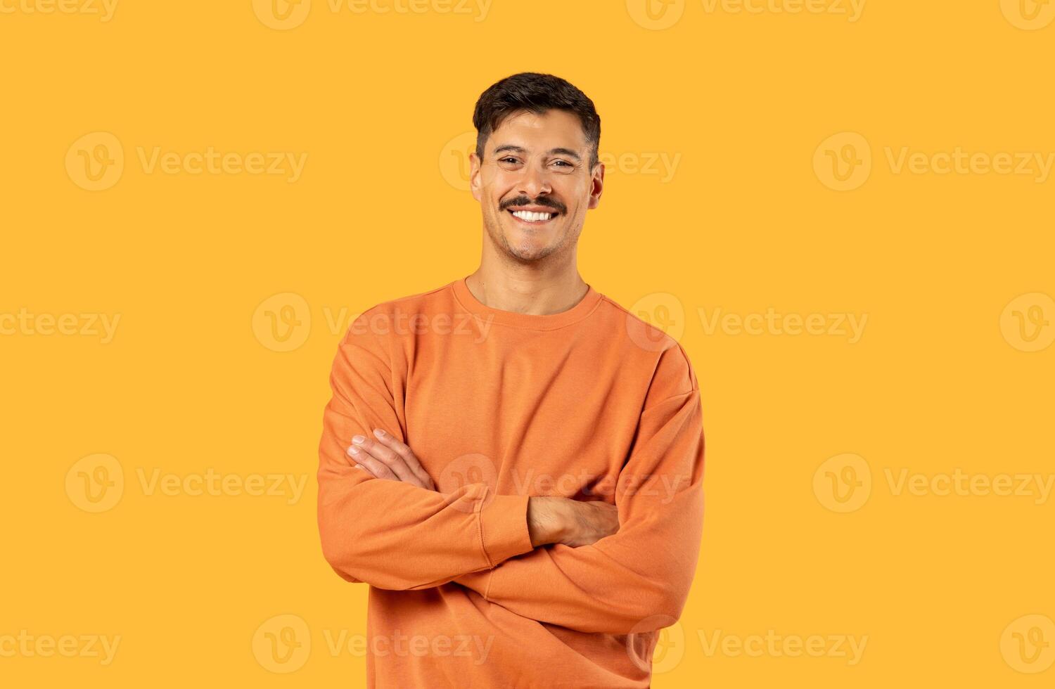 A cheerful man stands with arms confidently crossed, wearing a casual orange sweatshirt, against a solid yellow background photo