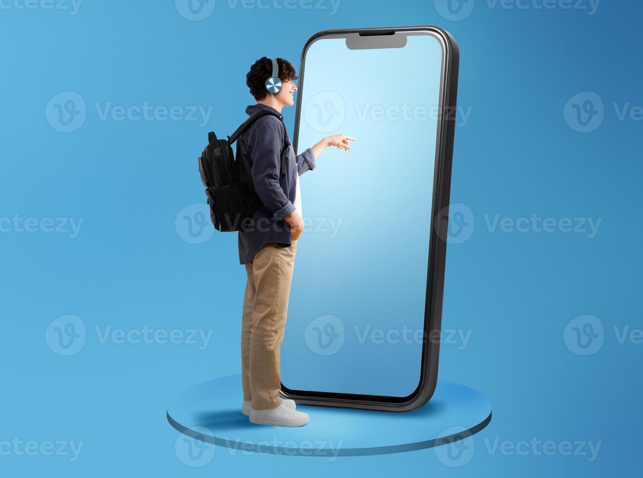 Side View Of Guy Using Large Cellphone Touching Screen Wearing Headphones Standing With Backpack Over Blue Studio Background. Panorama With Free Space For Text, Full Length photo