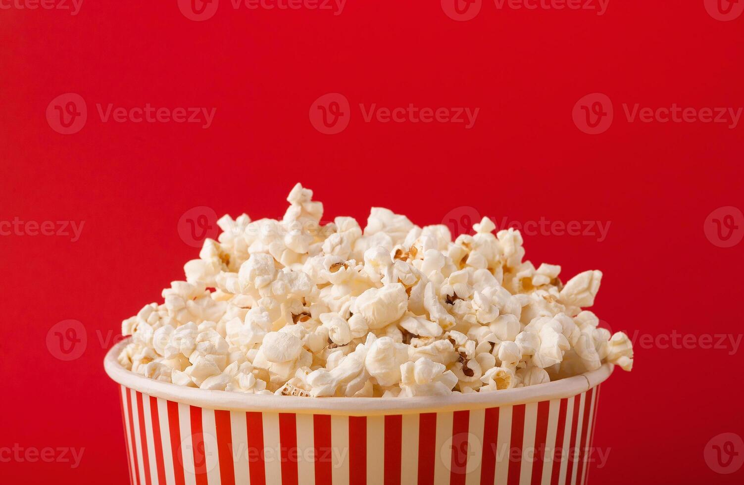 Popcorn in classic striped bukcet on red background. Fluffy maise paper box, copy space. Fast food and movie snack, entertainment concept photo