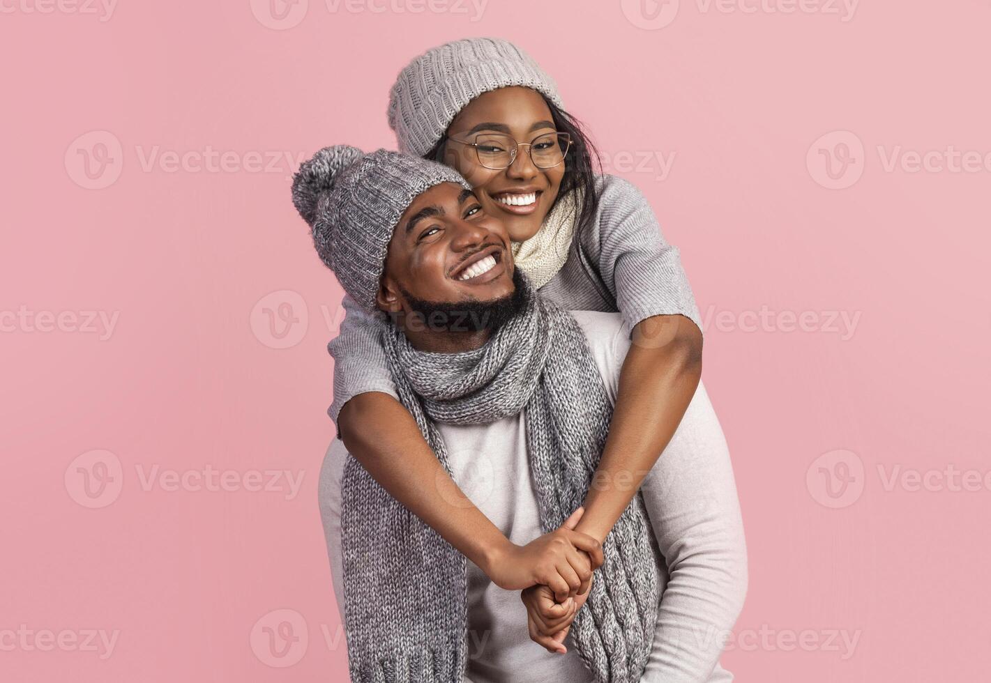 Playful black winter girl hugging her cheerful boyfriend from behind, pink background photo