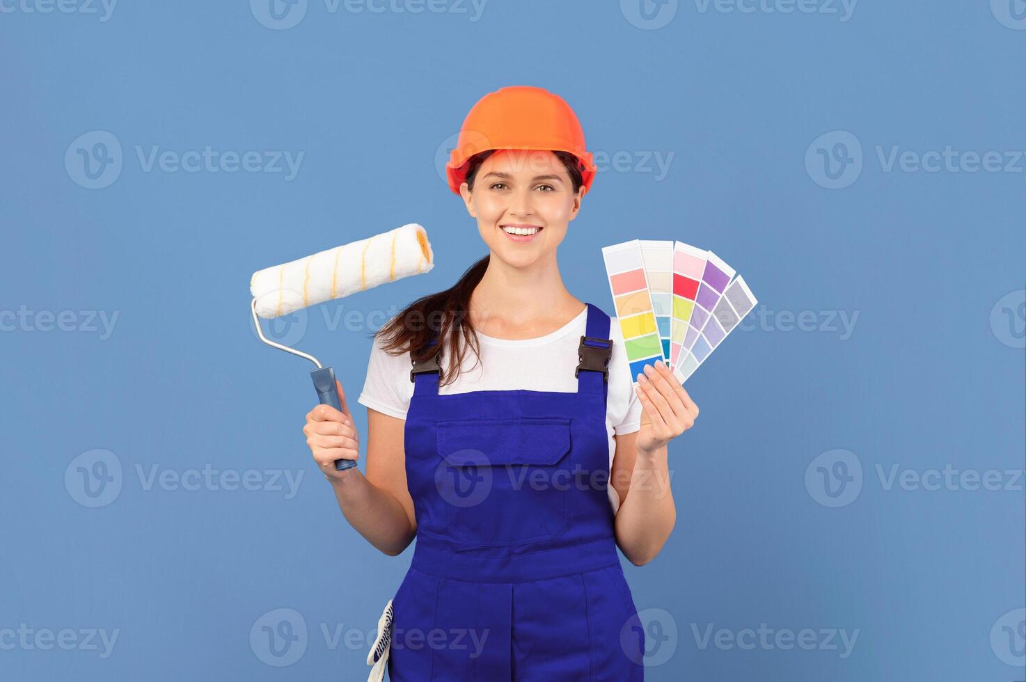 Young beautiful woman in coverall posing with color palette and roller paint brush over blue background, smiling painter lady in hard hat ready for house renovation or flat repairing, copy space photo