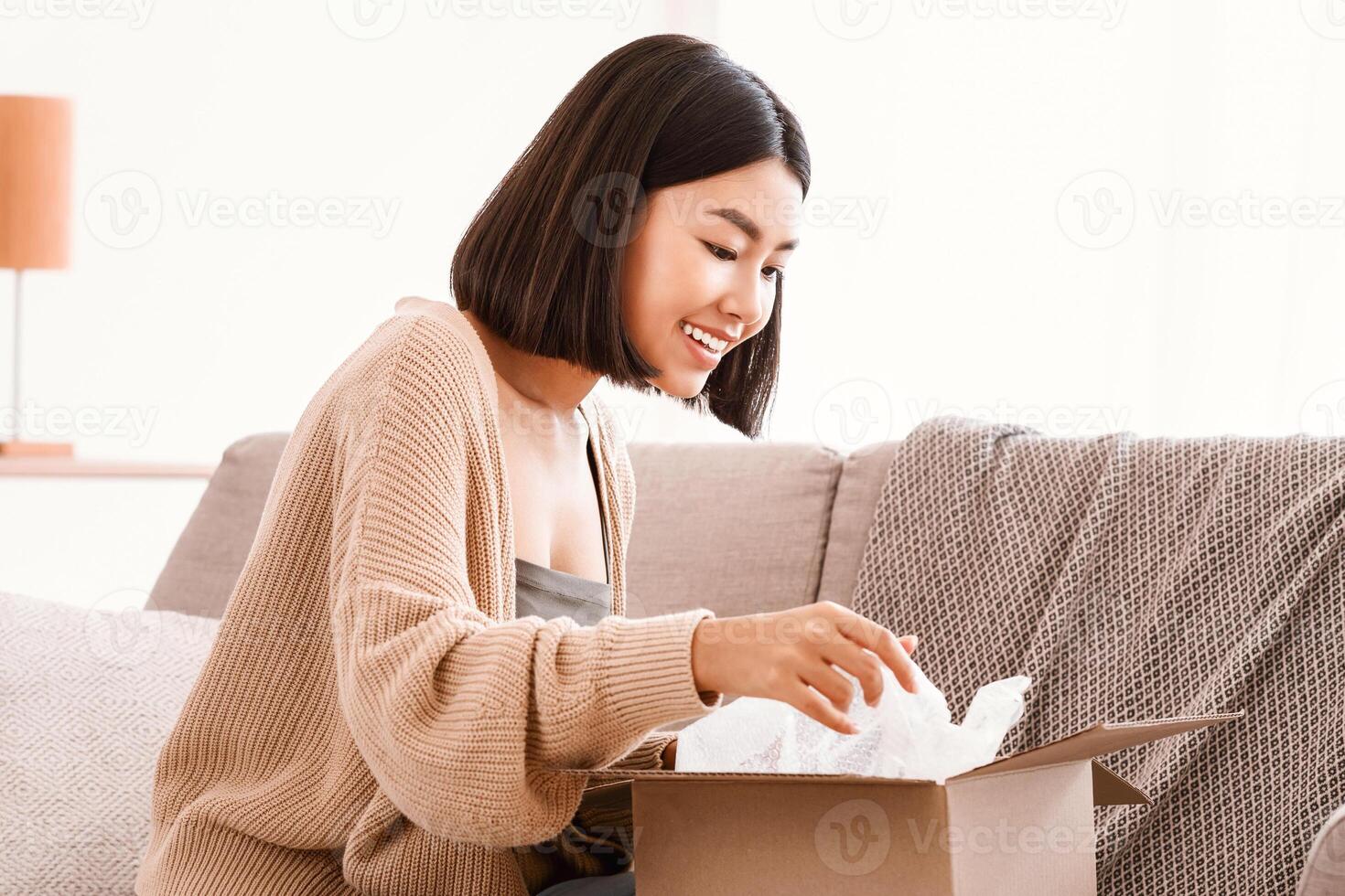 Fast Delivery Service And Satisfied Customer. Portrait of smiling curious asian female consumer opening and unpacking parcel cardboard box, happy woman sitting on the sofa in living room at home photo
