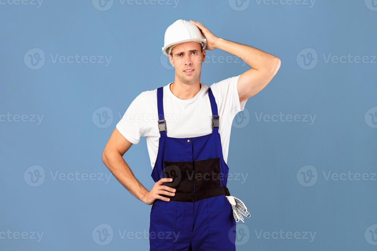 Confused handyman in protective helmet and coveralls touching head in frustration, male repair worker doesn't know how to start house renovation, standing over blue background, copy space photo