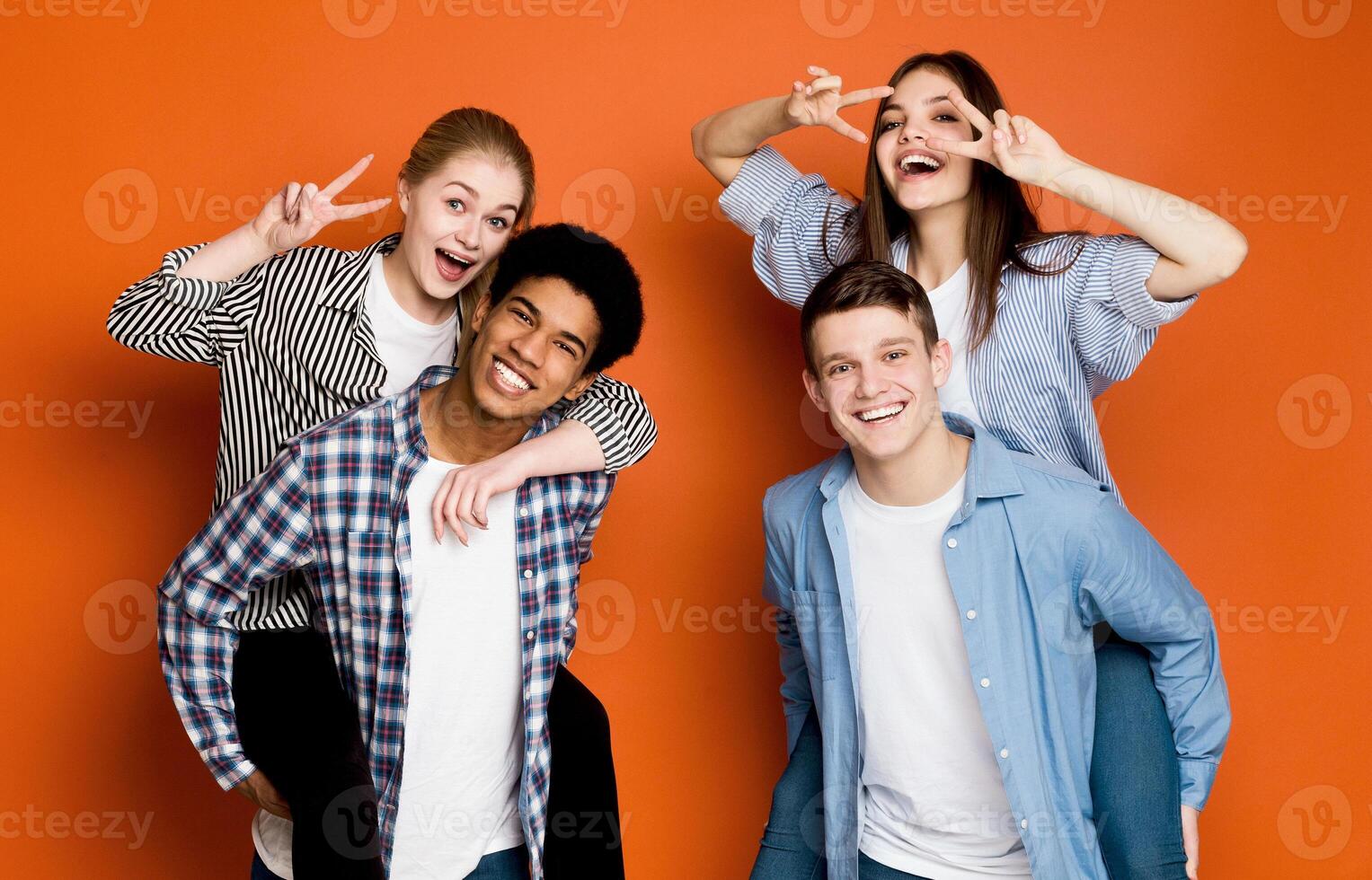 Happy friends having fun. Guys giving piggyback ride to girls, fooling over orange background photo