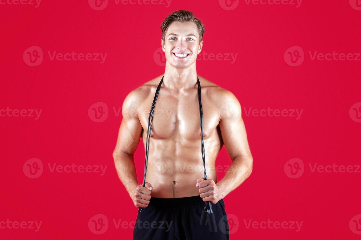 Handsome young sportsman with bare torso and jumping rope looking at camera and smiling on red studio background. Shirtless millennial guy ready for cardio workout or aerobic exercises photo