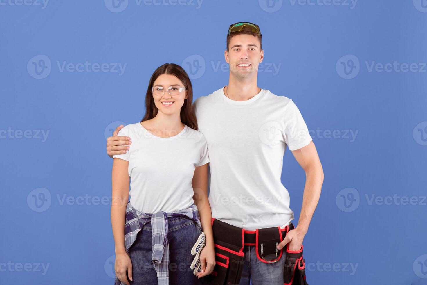 Repair Concept. Portrait Of Young Couple With Safety Glasses And Construction Belt Posing Together Over Blue Studio Background, Smiling Man And Woman Ready For Renovation And Home Makeover, Copy Space photo