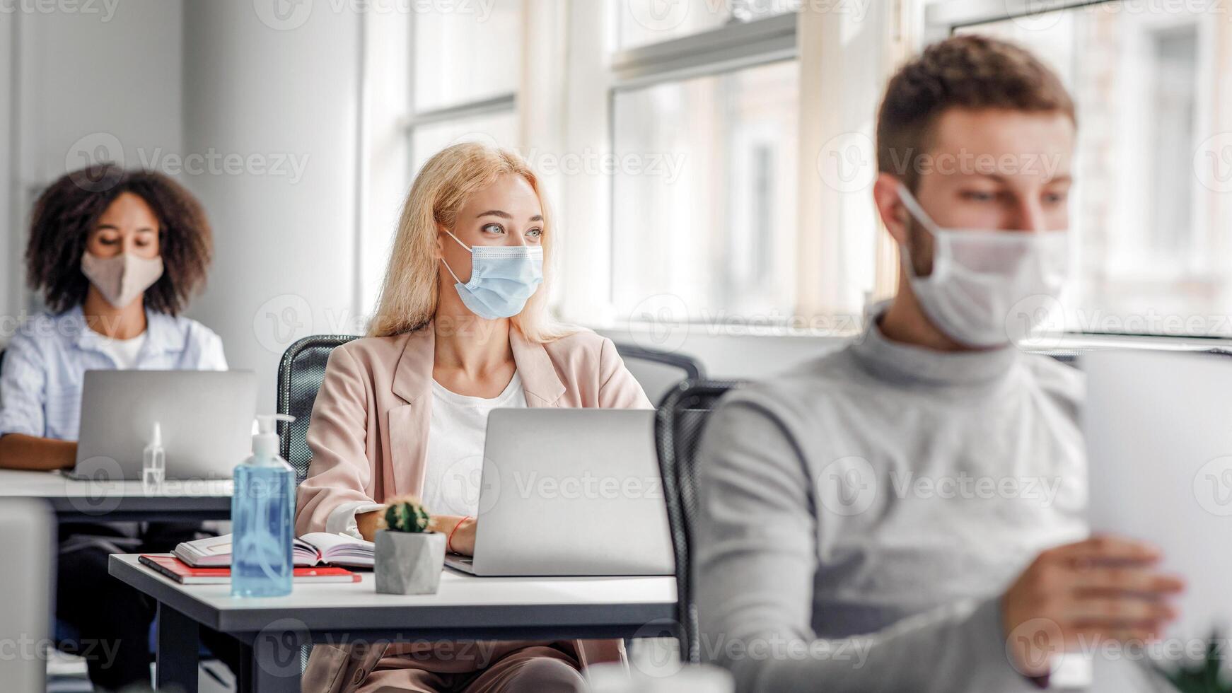 Teamwork on project. Multiracial employees in protective masks work on laptops at desks with antiseptics, focus on pensive blonde lady, looking out the window, coming up with idea, copy space photo