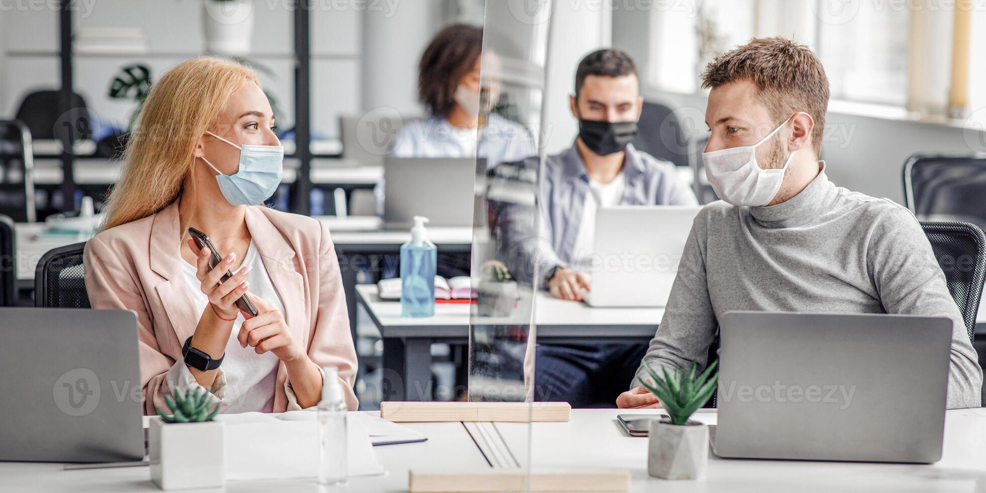Man and woman in protective masks maintain social distance at work. Employees discuss work sitting at table laptop with antiseptic, smartphone and protective glass in office interior, panorama photo
