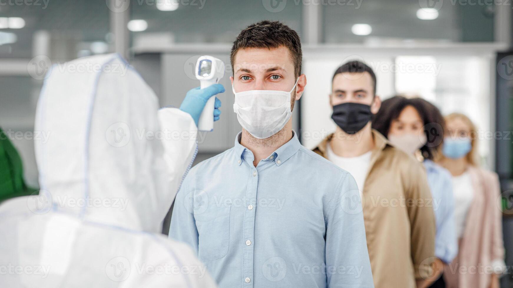 Modern technologies for determining temperature with non-contact thermometer. Man in hazmat suit measures temperature of multiracial colleagues in protective masks at entrance to office, free space, indoors photo